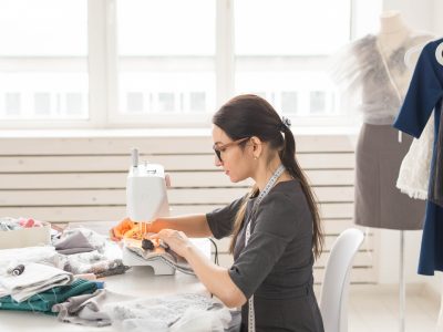 People and fashion concept - Young dressmaker woman sews clothes on sewing machine